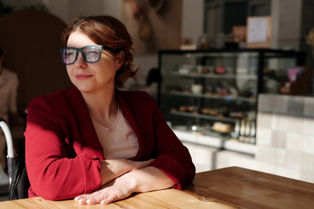 woman sitting at a restaurant thinking, looking to her right. she is wearing a red blouse and think black glasses. 